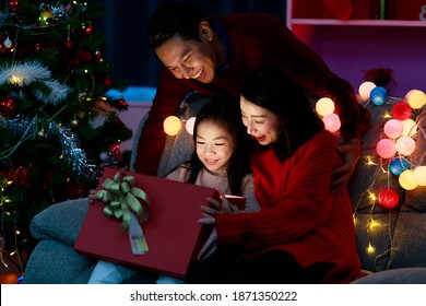 Cheerful Father, Mother And Daughter Opening And Holding Christmas Gift Box While Sitting On Sofa At Home. Happy Asian Family On Christmas Day. Merry Christmas And Happy Holidays. Christmas Holiday