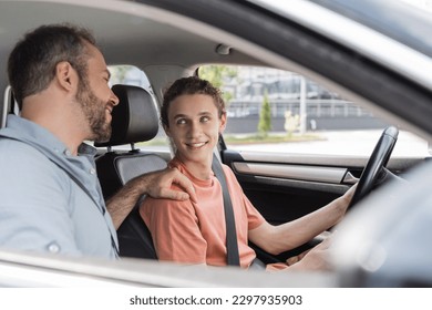 cheerful father putting hand on shoulder of teenage son while teaching him how to drive car - Powered by Shutterstock