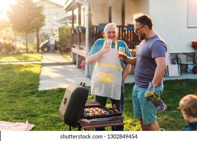 Cheerful Father And Son Drinking Beer While Standing Next To Grill In Backyard At Summer. Family Gathering Concept.