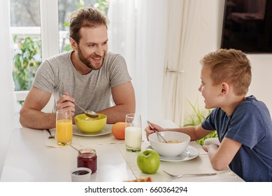 Cheerful father and son are eating cereals in morning. They are sitting at table in kitchen and smiling