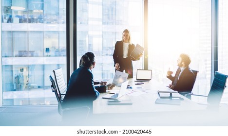 Cheerful female CEO in formal suit standing near table with laptop and explaining documents to group of coworkers during meeting in modern office - Shutterstock ID 2100420445
