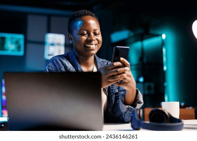 Cheerful female college student using mobile device to engage with friends on social media platforms. African american lady using cell phone to surf the web while sitting at desk in her home office. - Powered by Shutterstock