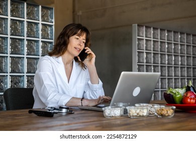 Cheerful female dietitian speaking on cellphone while working on netbook at table with various natural products in glass wood fashionable office. Low view, Horizontal. - Shutterstock ID 2254872129