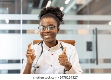 Cheerful female doctor giving a thumbs up while holding a jar of pills during a video call in a modern office setting. - Powered by Shutterstock