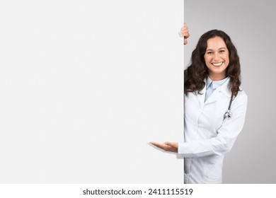Cheerful female doctor with stethoscope peeking out from behind blank white board, presenting space for text or graphics, grey background - Powered by Shutterstock