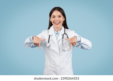 Cheerful female doctor with stethoscope pointing downwards with both hands, expressing excitement or highlighting something, set against clean blue background