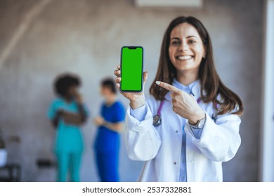 A cheerful female doctor in a white coat points to a smartphone with a green screen. Two other healthcare professionals in scrubs are visible in the blurred background. - Powered by Shutterstock