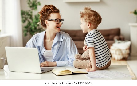 Cheerful female entrepreneur and little boy smiling and typing on laptop keyboard while sitting at table and working on freelance project at home together