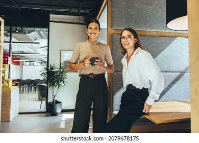 Cheerful Female Entrepreneurs Smiling At The Camera While Standing In A Modern Co-working Space. Two Happy Young Businesswomen Taking A Coffee Break While Working Together.