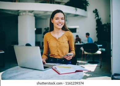 Cheerful female freelancer enjoying distance job sitting at cafeteria with laptop computer while waiting friend and checking notification on modern smartphone,сoncept of technology and communication