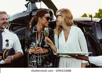 Cheerful Female Friends Having Wine Outside A Helicopter With Pilot Standing By. Women Enjoying Welcome Drinks By A Helicopter.