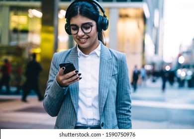 Cheerful female in glasses and headphones wearing stylish gray suit and using smartphone with joy while strolling down city street 