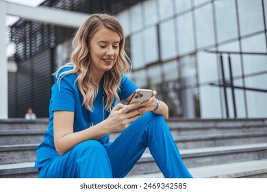 A cheerful female healthcare worker in blue scrubs sits on steps outside a building, browsing her smartphone during a break. - Powered by Shutterstock