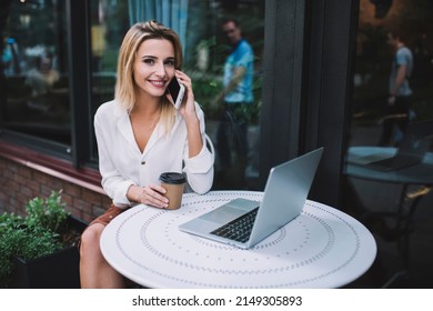 Cheerful Female Looking At Camera And Talking On Cellphone While Sitting At Table With Laptop And Takeaway Hot Drink In Outdoor Cafe