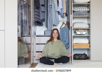 Cheerful Female In Lotus Position At Modern Wardrobe Storage. Happy Housewife Posing Meditating At Home Closet Full Of Clothing. Smiling Woman Relaxing After Domestic Cleaning Housework