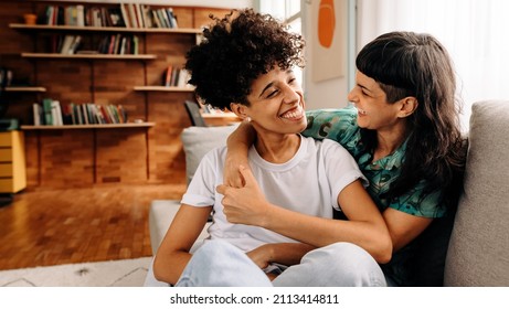 Cheerful Female Lovers Laughing Together. Happy Young Lesbian Couple Sharing A Laugh While Sitting Together In Their Living Room. Young Lesbian Couple Spending Quality Time Together.