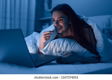 Cheerful Female Lying In Bed And Watching Movie On Laptop While Eating Popcorn In Dark Bedroom At Night