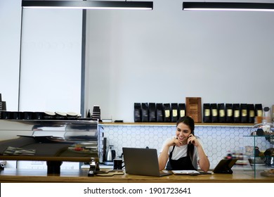 Cheerful female manager of coffee shop making smartphone call confirming ordering from clients online, smiling woman barista planning delivery talking on mobile phone at bar with devices and equipment
