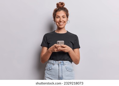 Cheerful female model uses mobile phone writes message on smartphone smiles toothily feels very happy dressed in casual black t shirt and jeans isolated over white background. Modern technologies - Powered by Shutterstock