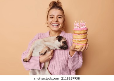 Cheerful female pet owner celebrates birthday of pug dog expresses love to animal smiles gladfully has good mood holds pile of delicious doughnuts with burning candles isolated over brown wall