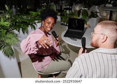 Cheerful female podcast host of African American Ethnicity playfully waving hand towards camera captured recording new episode with male guest at table in cafe with green plants, camera flash - Powered by Shutterstock