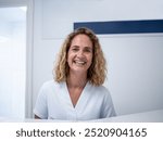 A cheerful female receptionist welcomes visitors in a bright, modern dental clinic. She has curly hair and wears a light-colored uniform, showcasing a friendly atmosphere.