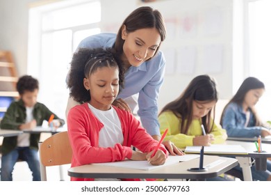 Cheerful female teacher helping girl with homework in classroom, black schoolgirl writing in notebook, sitting at desk with classmates on background - Powered by Shutterstock