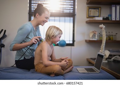 Cheerful female therapist while scanning shoulder of shirtless boy looking at laptop in hospital ward - Powered by Shutterstock