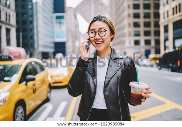 Cheerful female traveler in eyewear talking on\
mobile in roaming walking on avenue of big city with coffee to go,\
asian girl calling taxi contact on mobile service operator\
strolling in downtown