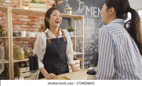 cheerful female waiter taking order from young female costumer standing at cafe counter. two laughing women friends meeting in coffee shop while one working as waitress. happy regular client indoors