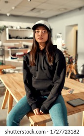 Cheerful female worker looking at camera while posing in custom apparel, baseball cap and hoodie. Young woman working at custom T-shirt, clothing printing company. Vertical shot