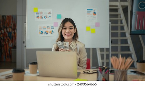 A cheerful financial advisor proudly holds stacks of dollar bills illustrating success and financial growth. Charts and graphs on the whiteboard highlight business performance and financial planning - Powered by Shutterstock