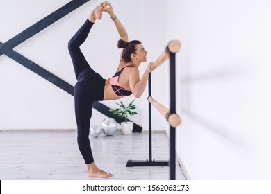 Cheerful fit woman standing at wooden barre raising leg up while stretching and doing split in light studio laughing happily - Powered by Shutterstock