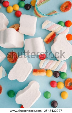 A cheerful flat lay composition of assorted colorful candies and marshmallows arranged on a light blue background. Featuring rainbow ribbon candy, white marshmallow clouds, colorful chewy balls.