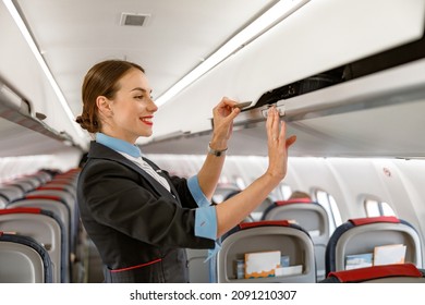 Cheerful flight attendant closing overhead luggage bin in airplane - Shutterstock ID 2091210307