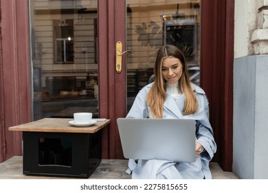 cheerful freelancer in blue trench coat using laptop while sitting next to cup of coffee in outdoor cafe in Vienna - Powered by Shutterstock