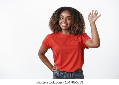 Cheerful, Friendly African American Woman With Afro Hairstyle, Raising Hand And Waving Camera With Pleasant Smile As Saying Hi, Hello Or Goodbye, Greeting You, White Background