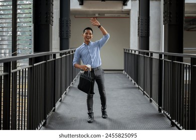 A cheerful, friendly Asian millennial businessman waving to greet his colleague while walking along the building corridor, holding a coffee cup and a briefcase. businesspeople concept - Powered by Shutterstock