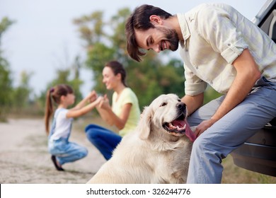 Cheerful Friendly Family Is Resting In Park. The Man Is Playing With Dog And Leaning On Car. The Mother And Daughter Are Kneeling And Holding Hands. They Are Smiling