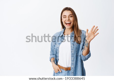 Cheerful friendly girl saying hello, winking and smiling, waving hand at camera, say hi and greet you, standing against white background