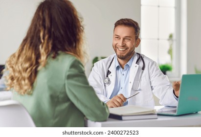 Cheerful friendly male doctor at medical office in hospital, showing examination results on laptop monitor screen and having consultation with young woman patient sitting in clinic. - Powered by Shutterstock