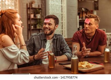 Cheerful friends. Happy cheerful contended smiling beaming joyous nice-looking vigorous young-adult three friends sharing a laugh at the bar and playing a hedbanz together