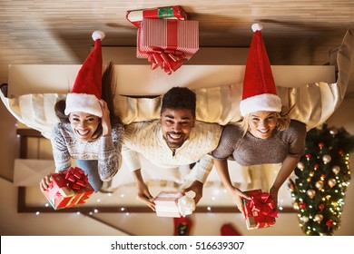 Cheerful friends holding Christmas gifts in their hands and lying upside down on bed.