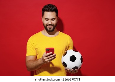 Cheerful fun young bearded man football fan in yellow t-shirt cheer up support favorite team hold soccer ball use mobile cell phone read text chat isolated on plain dark red background studio portrait - Powered by Shutterstock