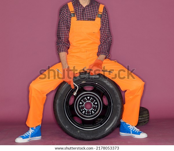 cheerful funny auto mechanic with wheel\
and tools.technician in orange overalls on lilac background.auto\
business car service\
concept.isolated.