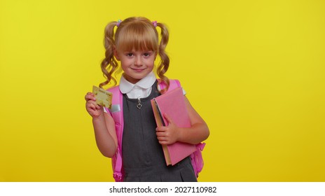 Cheerful Funny Blond Teenage Schoolgirl Kid In Uniform Hold In Hand Bank Credit Card Showing Thumbs Up Gesture, Purchases Online Shopping. Yellow Studio Background. Teenage Education, Back To School