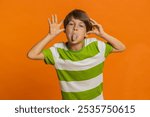 Cheerful funny bully young cute boy showing tongue making faces at camera, fooling around, joking, aping with silly face, teasing. Preteen child kid 6 years old on orange background. Little children