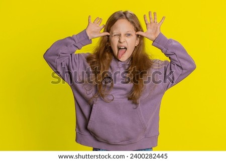 Cheerful funny bully young cute girl showing tongue making faces at camera, fooling around, joking, aping with silly face, teasing. Preteen redhead child kid on yellow background. Little children