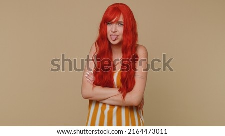 Cheerful funny ginger woman in tank top showing tongue making faces at camera, fooling around, joking, aping with silly face, teasing. Young redhead girl isolated alone on beige studio wall background