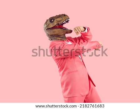 Cheerful funny and humorous man in dinosaur rubber mask dances isolated on pastel pink background. Young man in formal pink wears absurd animal mask on his head. Creative concept for advertising,